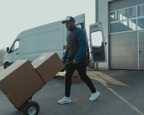 Courier pushing a trolley with boxes near a van at a warehouse, showcasing delivery services.